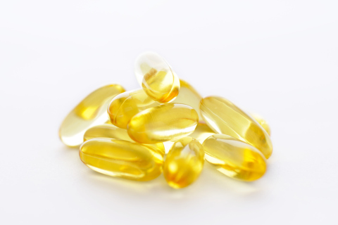 A pile of yellow transparent gel capsules, infused with vitamin E oil, are scattered against a plain white background. The capsules have a glossy surface that reflects light, highlighting their smooth, oval shape.