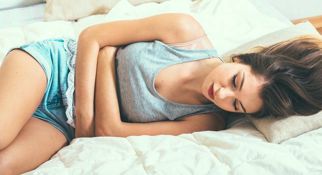 A woman lies on her side on a bed, clutching her stomach with both hands, clearly experiencing menstrual cramps. She has long hair, is wearing a grey tank top and blue shorts, and is in visible discomfort. The bed has white sheets, and she rests her head on a pillow as part of the Added Benefits Series.