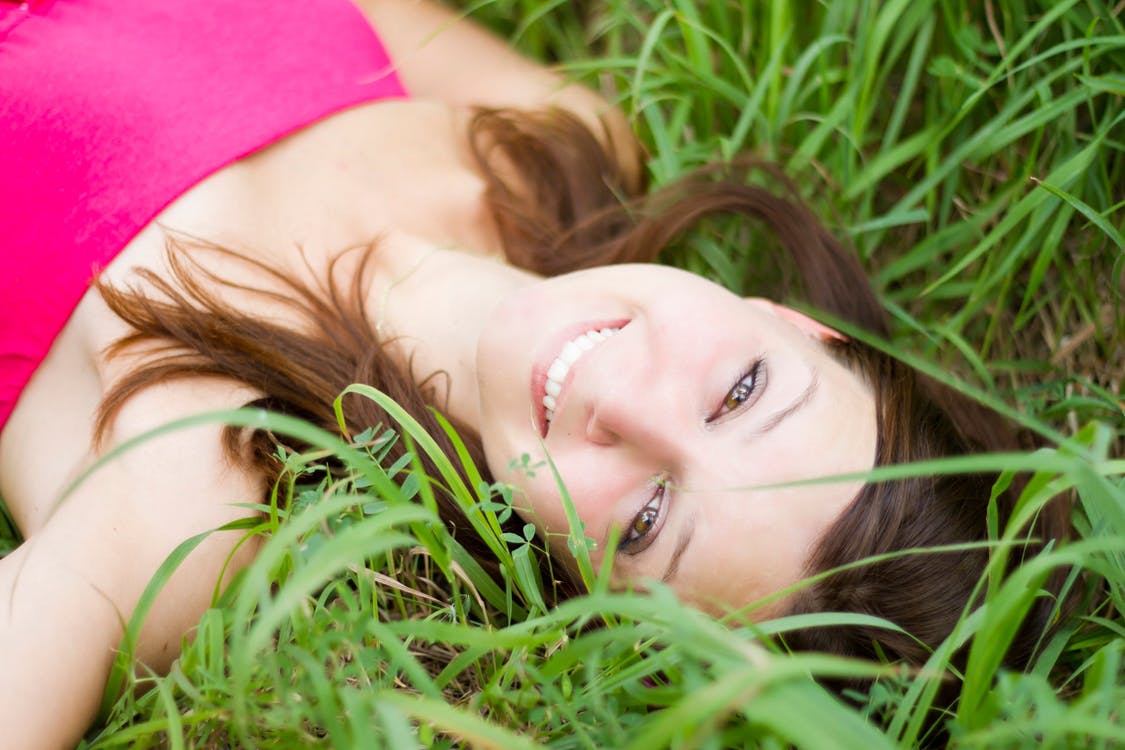 A woman is lying in a grassy field, looking up at the camera with a smile. She has long brown hair and is wearing a pink top. The green grass surrounds her, creating a natural and serene setting—perfect for discovering 5 Steps To A Happier You.