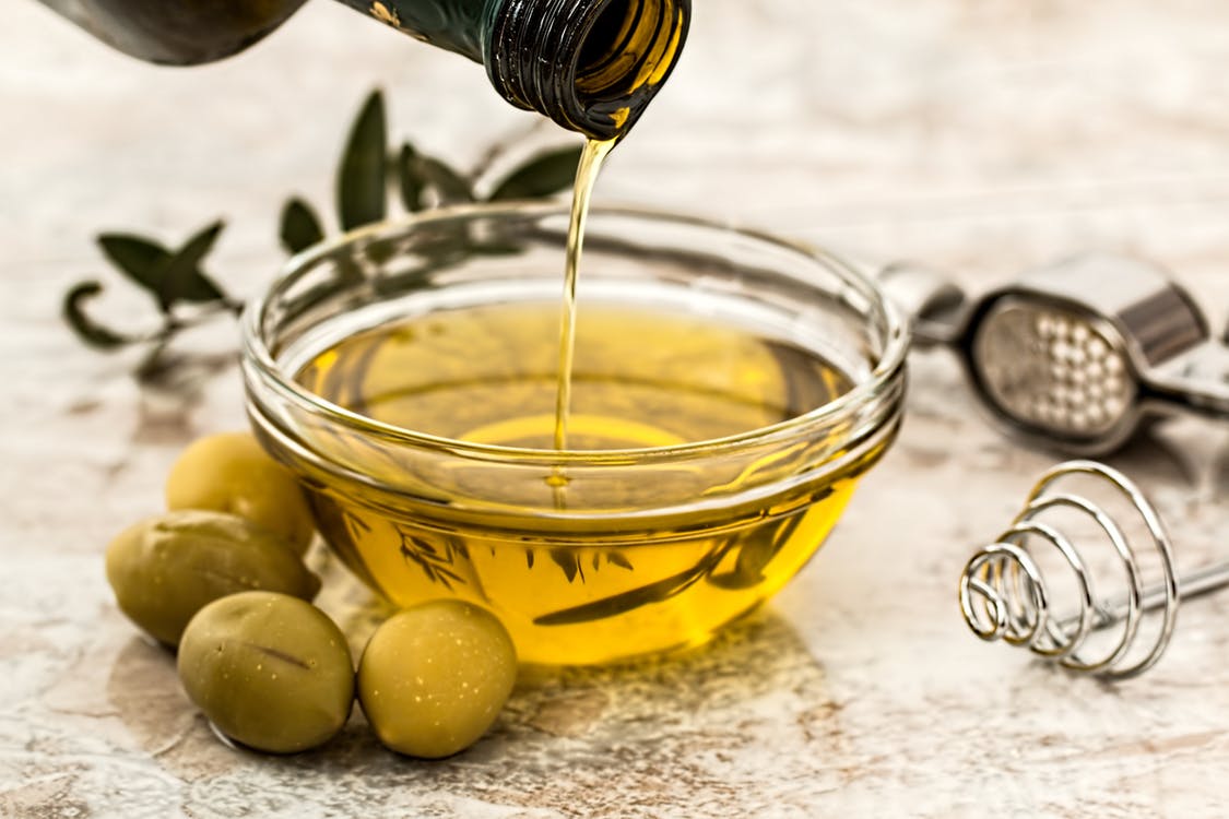 A stream of olive oil is being poured from a bottle into a clear glass bowl filled with glx3 ingredients. Beside the bowl, there are three whole green olives and kitchen utensils, including a whisk and a garlic press, set on a textured surface.