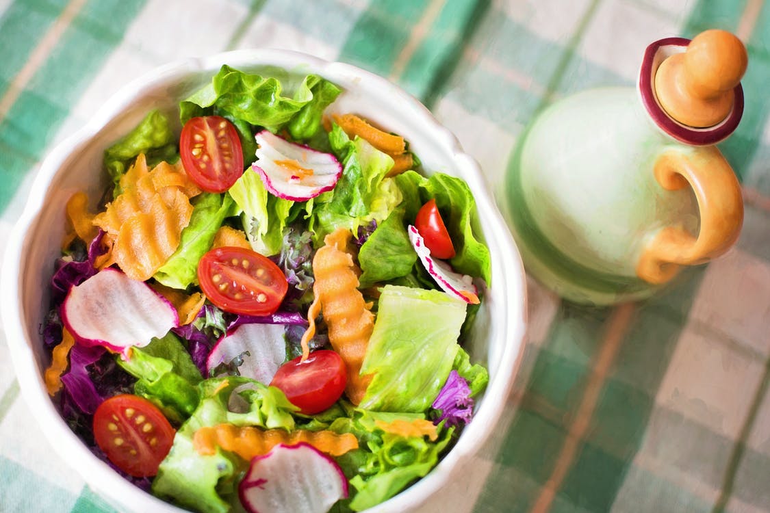 A fresh salad in a white bowl containing lettuce, cherry tomatoes, radish slices, and crinkle-cut carrot pieces. The bowl is placed on a green and white checkered tablecloth. Complementing this vibrant meal is a decorative vinegar or oil bottle to the right—ideal for those on a plant-based diet supplement regime.