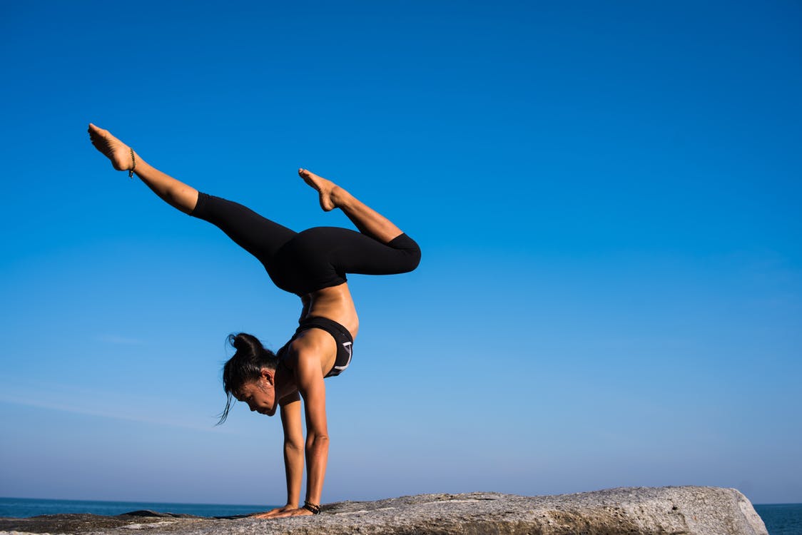A woman wearing black athletic wear performs a handstand on a rocky surface against a clear blue sky. One leg is extended vertically while the other is bent, showcasing her balance, strength, and dedication to bone health. The background includes a faint horizon line with a hint of the sea.