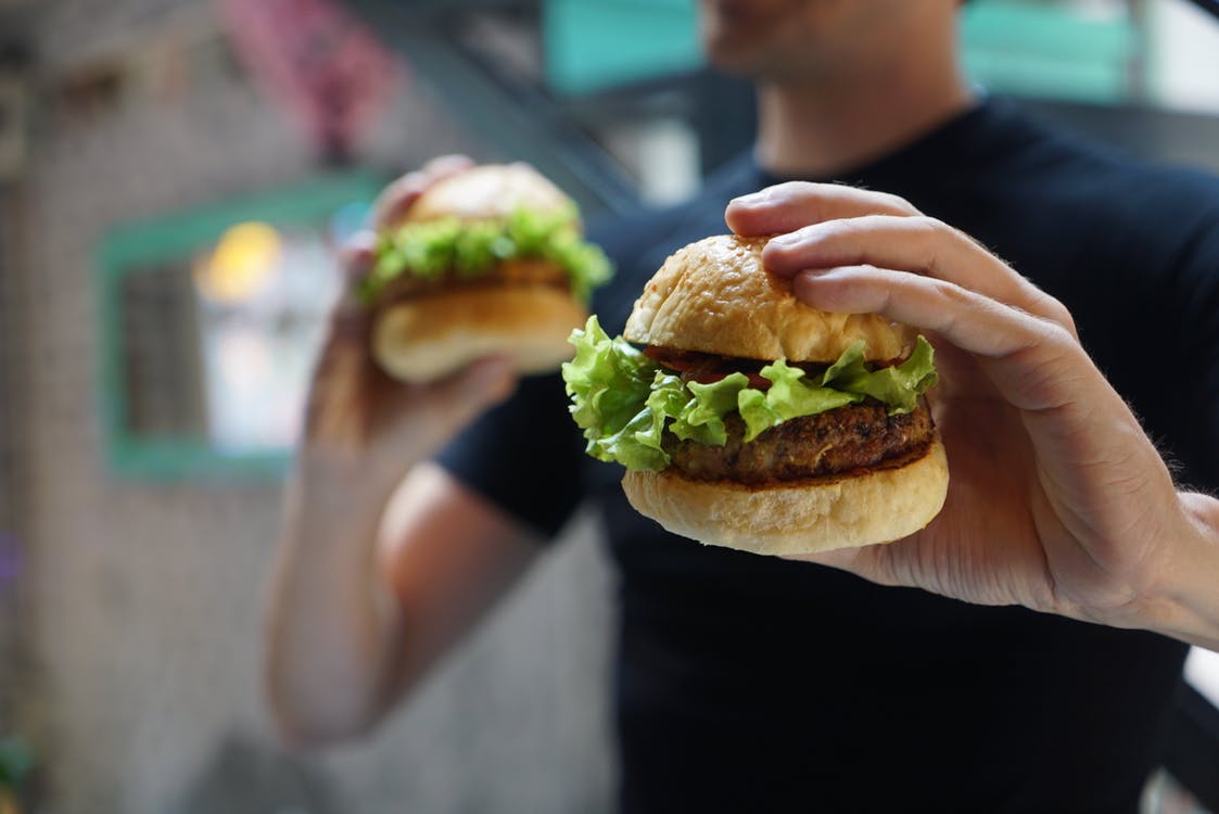 A person in a black shirt holds two burgers, each with lettuce and a patty, in both hands. The background is blurred, focusing on the burgers. The scene appears to be in an outdoor or casual dining setting.