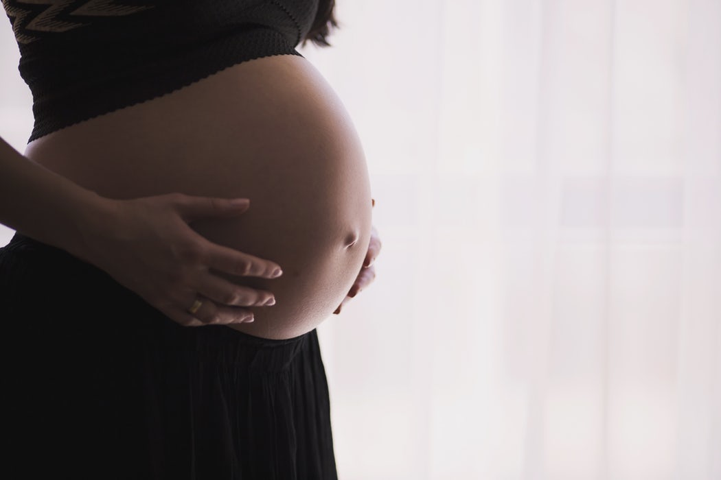 A pregnant woman gently cradles her belly with both hands. She is wearing a black skirt and a dark top. The background is softly lit with sheer white curtains, creating a serene and peaceful atmosphere, perhaps reflecting on the omega-3 benefits for fertility that helped her journey.