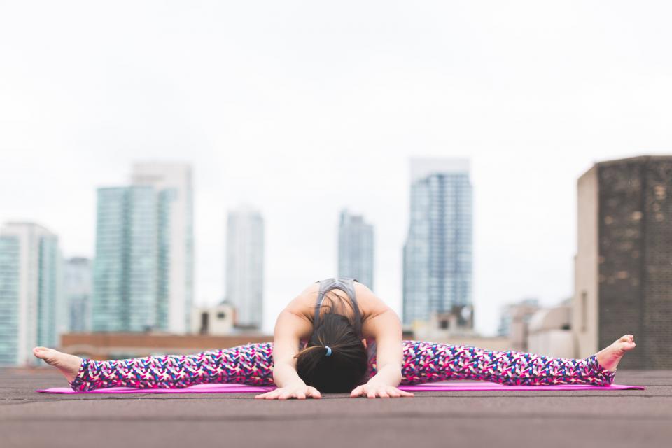 Person performing a wide-angle seated forward bend yoga pose on a pink mat. They are on an outdoor rooftop with tall city buildings in the background, showcasing how yoga can be practiced anywhere. The person, dressed in colorful, patterned leggings and a tank top, exemplifies why many ask, "Is Yoga Good for Arthritis?