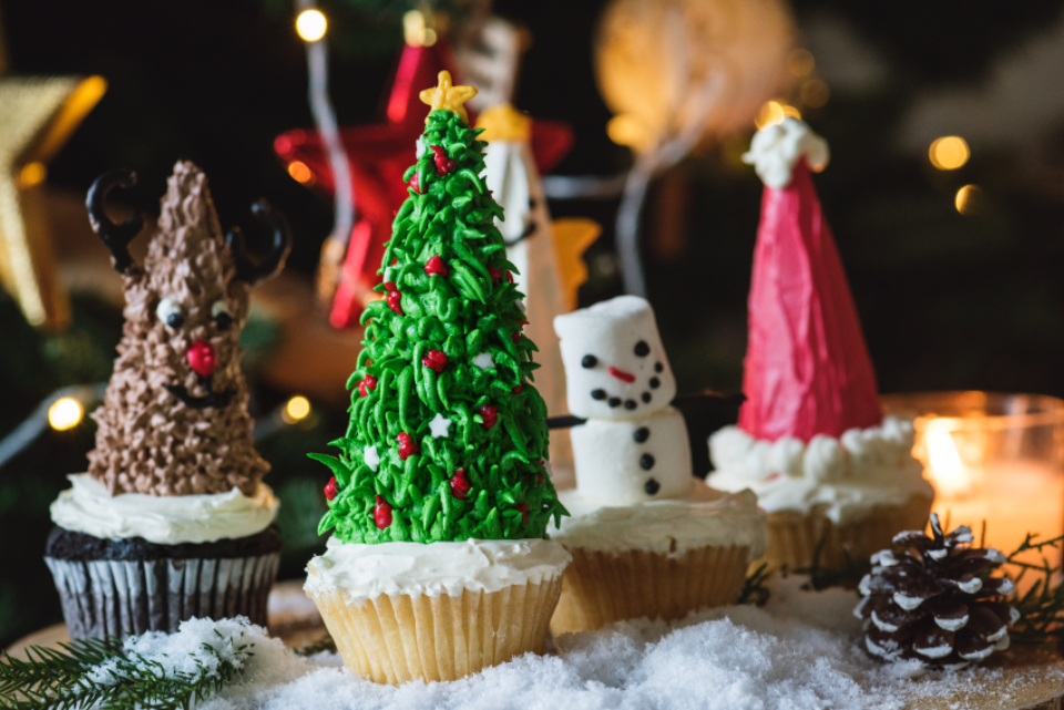 Festive cupcakes decorated for the holiday season. A tree-shaped cupcake with green icing and red ornaments, a reindeer with chocolate antlers, a snowman with a carrot nose, and a Santa hat. Surrounded by snow-like icing, pinecones, and warm glowing lights—perfect to incorporate health plan tricks for the holidays!