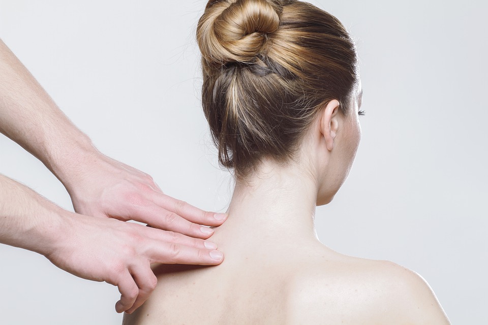 A person with blonde hair in a bun is receiving one of the best massages for arthritis on the upper back and neck from another individual against a plain, light gray background. The person getting the massage is unclothed on the upper back and shoulders.