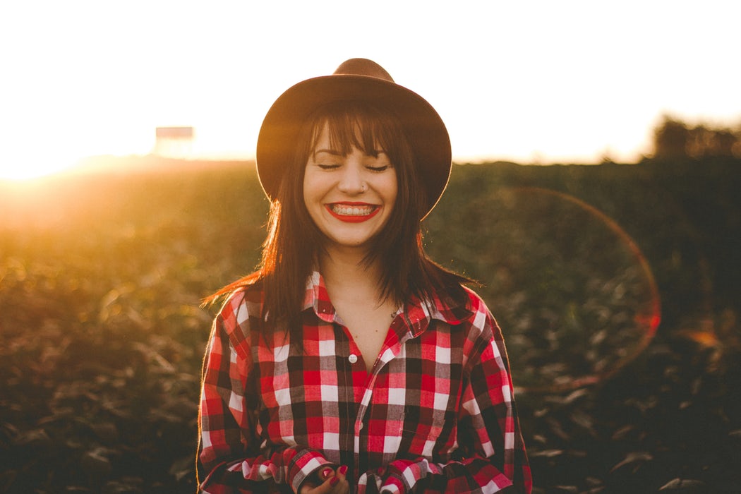 A woman with long dark hair and red lipstick stands in a field at sunset, smiling with her eyes closed. She wears a black hat and a red and white plaid shirt, embodying the tranquility often highlighted in the Added Benefits Series: Nervous System. The backdrop blends green fields with golden sunlight.