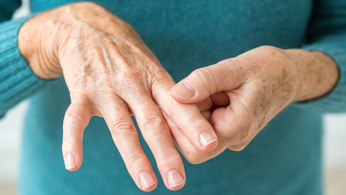 Close-up of a pair of elderly hands with visibly wrinkled and freckled skin. The fingers of one hand gently press the knuckles of the other, reminiscent of the care often discussed in "5 Juvenile Arthritis Facts You Never Knew." The person is wearing a teal long-sleeved shirt, shown against a neutral background.