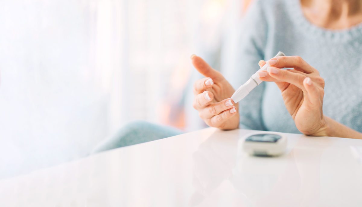 A person with diabetes is holding a blood sugar test strip and pricking their finger. A glucometer lies on the white surface in front of them. The background is bright, suggesting a well-lit room.