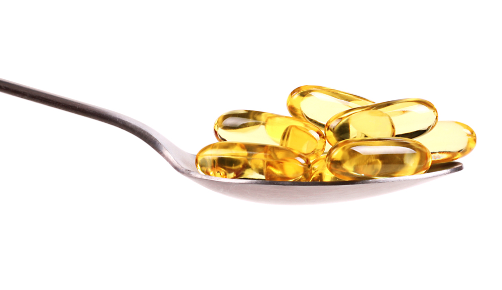 A metal spoon holds several glossy, yellow gel capsules filled with liquid against a plain white background. The capsules, often associated with omega-3 supplements, are oval and translucent, allowing the light to reflect off their shiny surfaces.