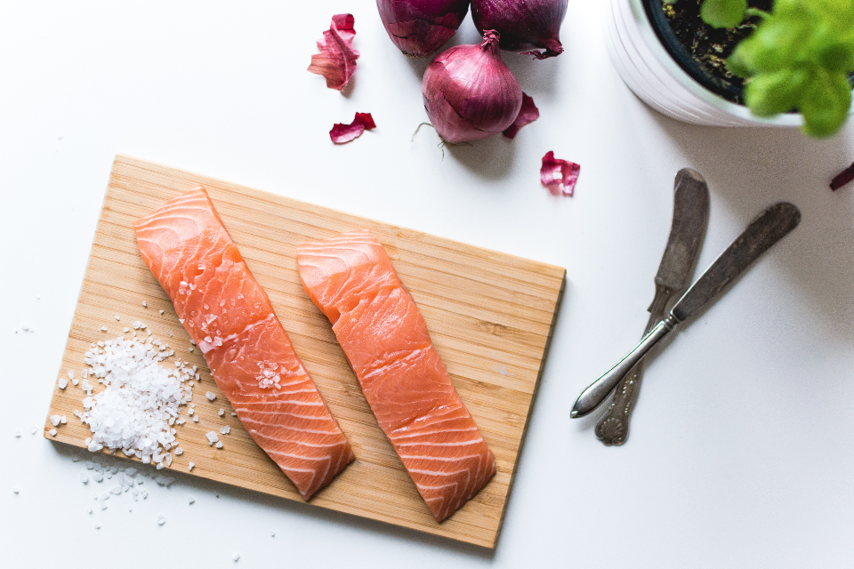 Two raw salmon fillets, rich in omega-3, rest on a wooden cutting board with a small pile of coarse sea salt beside them. In the background, three red onions, a potted plant, and two vintage utensils sit gracefully on a white surface.