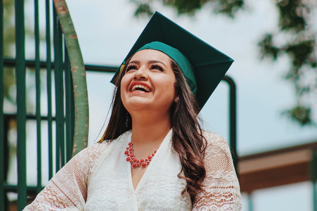 A woman in a green graduation cap and white lace dress smiles brightly while looking up, celebrating the added benefits series of her achievements. She is wearing a red necklace and standing against a green railing with trees in the background.