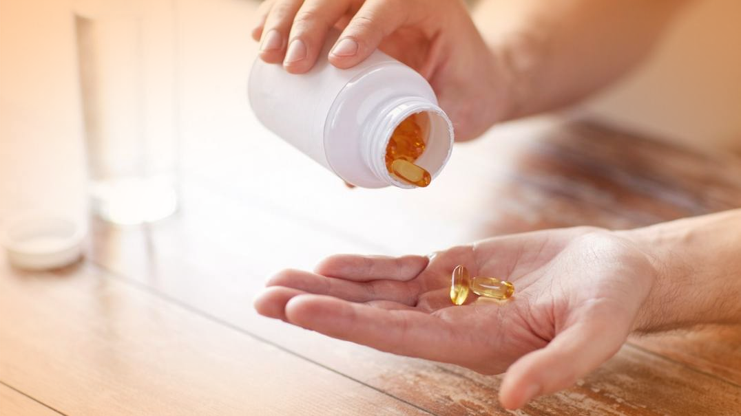 A person is holding and pouring yellow capsule pills from a white bottle into their hand. The scene is set on a wooden table with a glass of water in the background, suggesting they might be considering Omega-3 for health benefits or reflecting on cholesterol levels' impact on longevity.