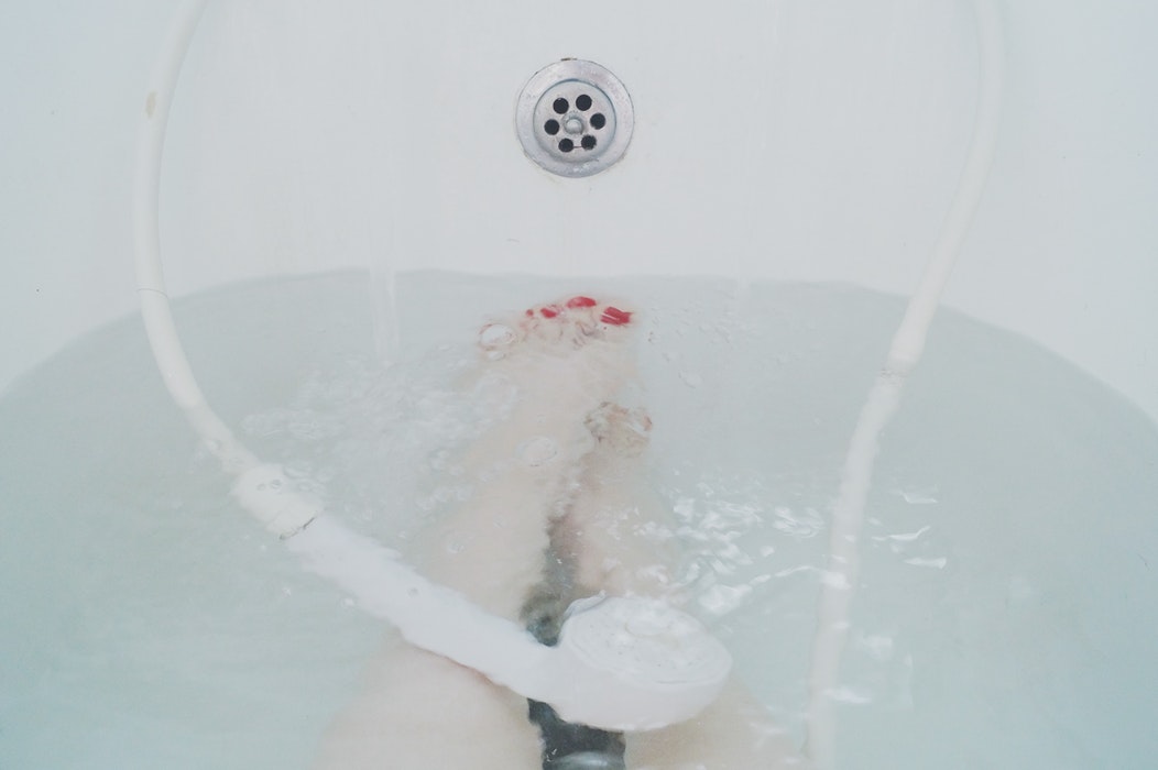 A person is relaxing in a bathtub with water at mid-level. Only their legs and feet are visible, and they are slightly submerged. A handheld showerhead rests on the side of the tub. Bubbles can be seen in the soapy water surrounding their legs, providing a soothing heat treatment for arthritis relief.