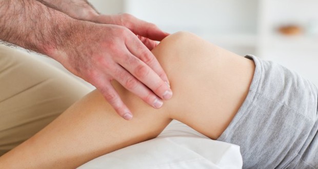 A close-up of a person's hands gently massaging a knee, perhaps suggesting to start using cold for arthritis pain relief. The person on the receiving end is lying down in light-colored shorts, with the background blurred to focus attention on the knee and hands.