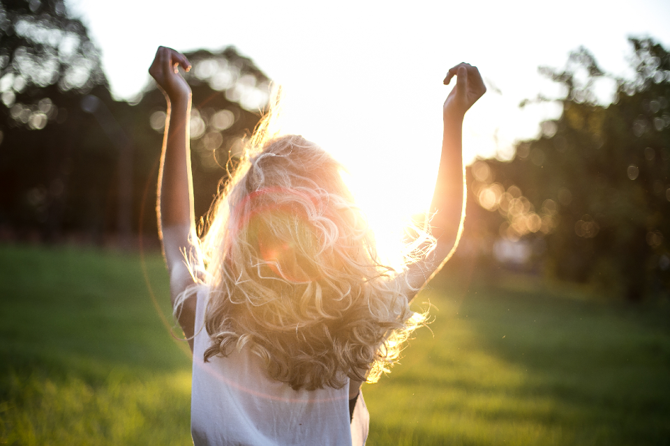 A person with long, wavy hair stands outside with their arms raised towards the sky, facing the sun. The scene is bright, sunlight streaming through their hair. Amidst green grass and trees, they embody natural vitality, much like the omega-3 benefits for anti-aging enhances one's youthful glow.