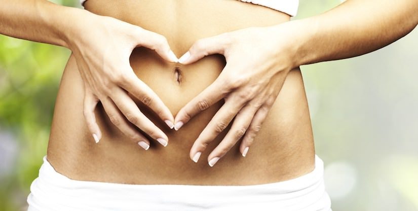 A person’s bare midriff is shown with both hands forming a heart shape around their navel, illustrating the Added Benefits Series: How to Improve Gut Health Naturally. The background is blurred with green and yellow colors, while the individual wears white clothing below the waist.