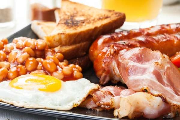 A plate of breakfast food featuring crispy toast, baked beans, an over-easy egg, and slices of bacon—commonly found on lists of foods that cause inflammation. In the background, there's a blurred glass of orange juice. The plate rests on a light-colored table surface.