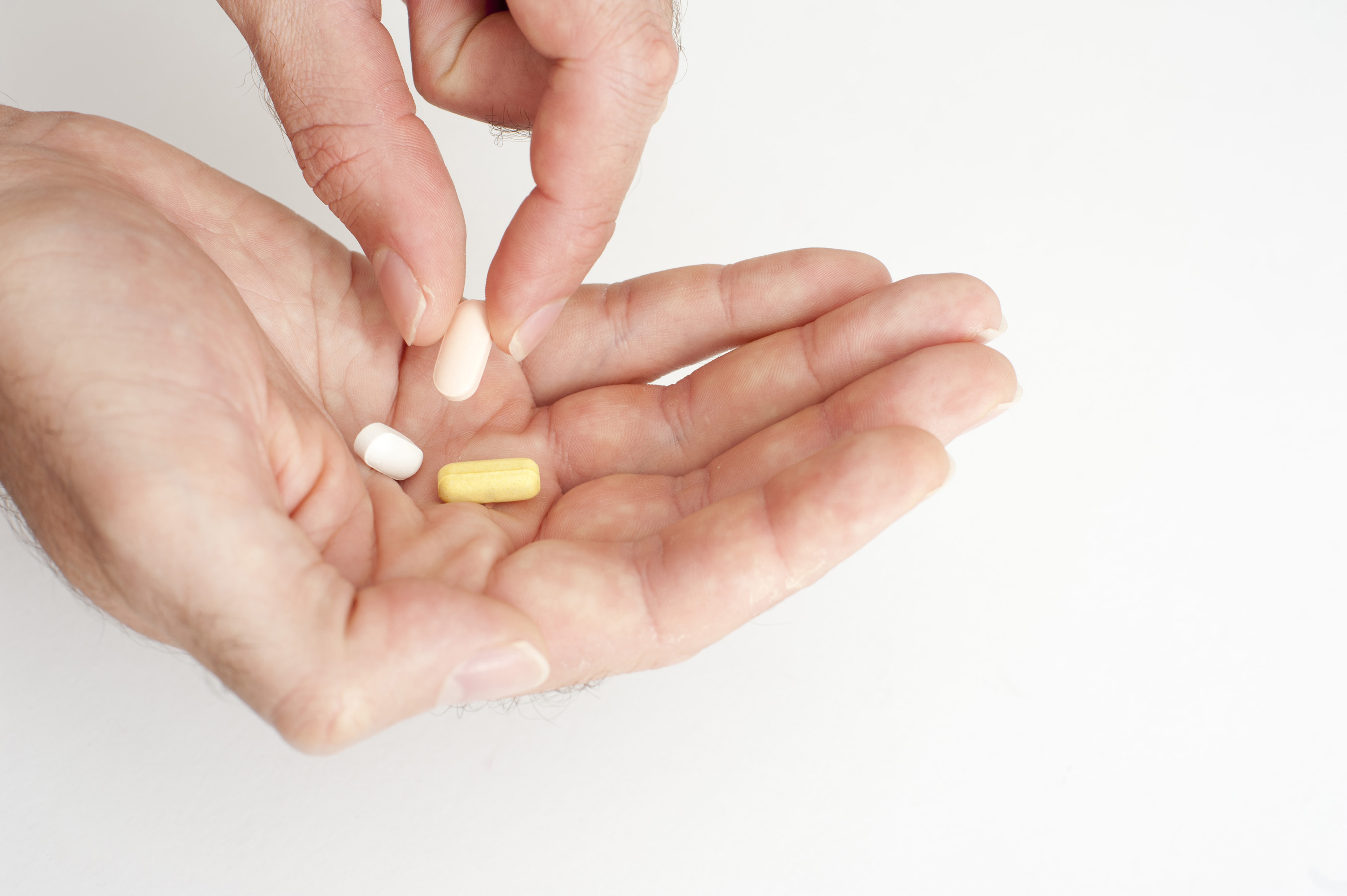 Close-up of hands holding various pills, likely vitamins for joint pain relief, against a plain white background. One hand is cupped, holding three pills: one white, one yellow, and one oblong pinkish-white. The other hand is picking up the pinkish-white pill with two fingers.