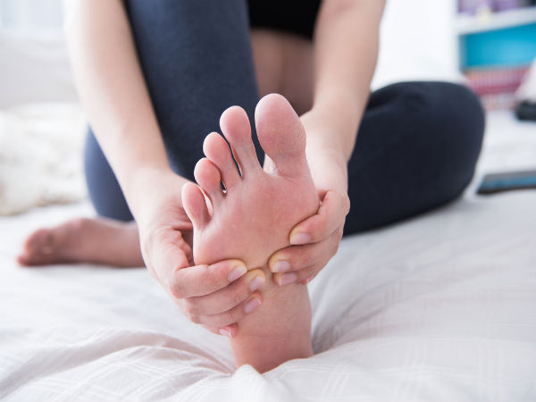 A person sitting on a bed is shown massaging the sole of their bare foot with both hands, indicating they might be dealing with gout signs symptoms and treatments you can’t ignore. The foot is prominently displayed, and the background is softly blurred, creating a cozy and relaxed indoor environment.