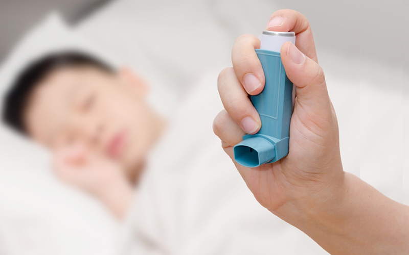 A hand holding a blue inhaler in the foreground, with a blurred background showing a person lying down, possibly sleeping, on a bed. The focus is on the inhaler, highlighting its importance for respiratory issues. This image complements discussions on 4 natural ways to fight bronchial asthma symptoms for adults.