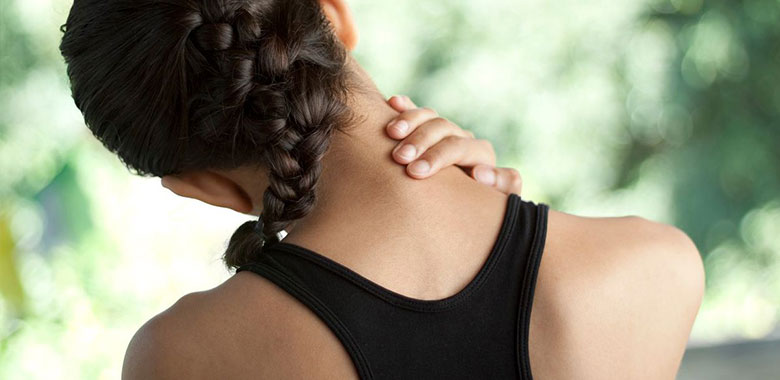 A person with a braid is seen from the back, wearing a black sleeveless top. They are holding the back of their neck with their left hand, suggesting discomfort or thoracic spine pain symptoms. The background is blurred greenery.