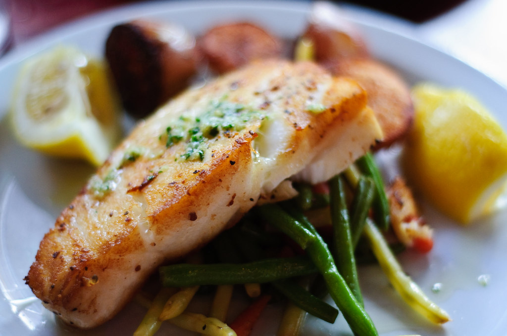 Plate of seared white fish garnished with chopped herbs, served over a bed of sautéed green beans and mixed vegetables. The dish is accompanied by lemon wedges and golden-brown roasted potatoes.