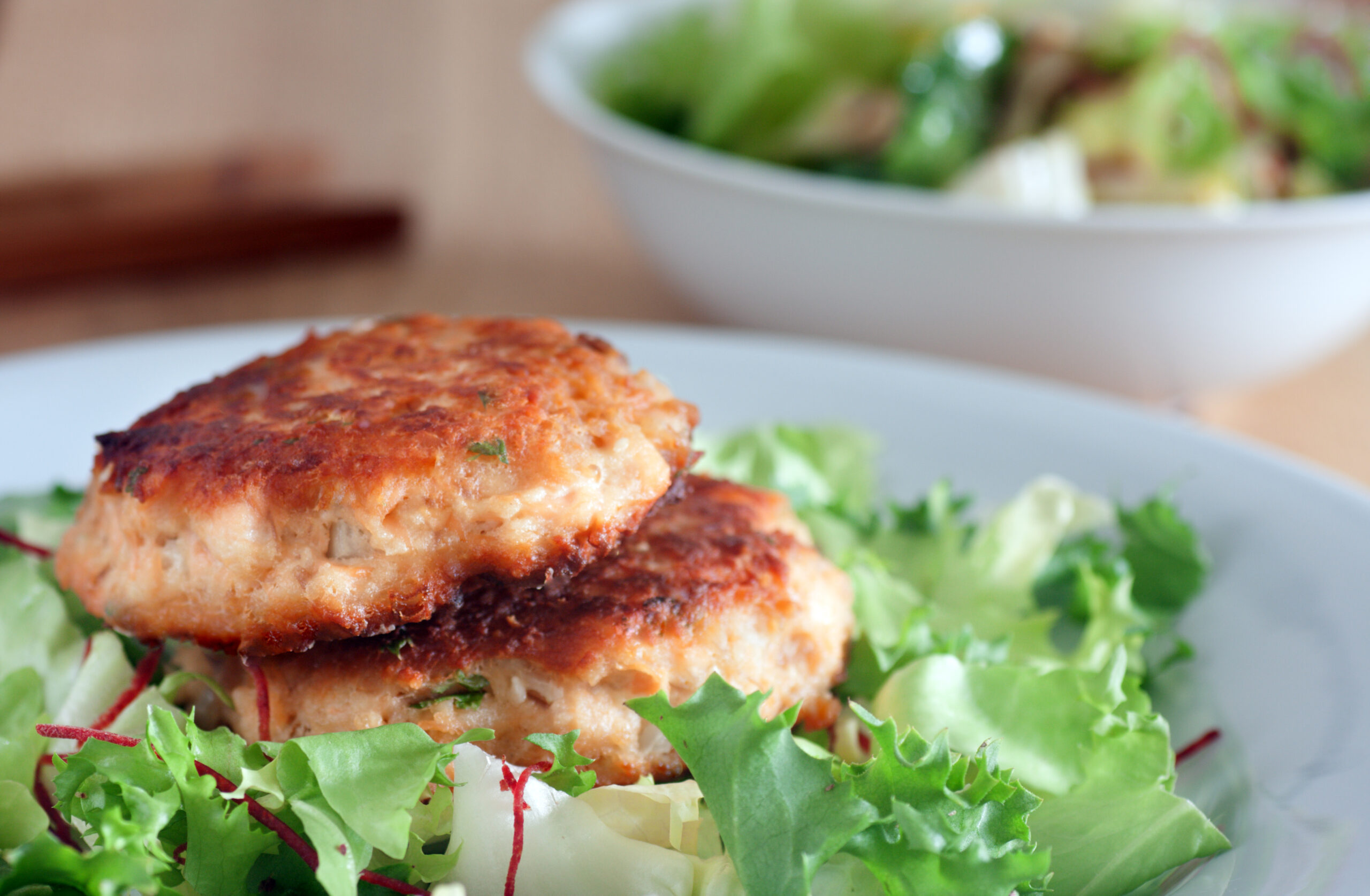 Two golden-brown salmon patties rest on a bed of fresh mixed greens, featuring lettuce, beet strips, and herbs. In the background, there is a white bowl filled with a green salad, slightly out of focus. The setting is on a wooden table.