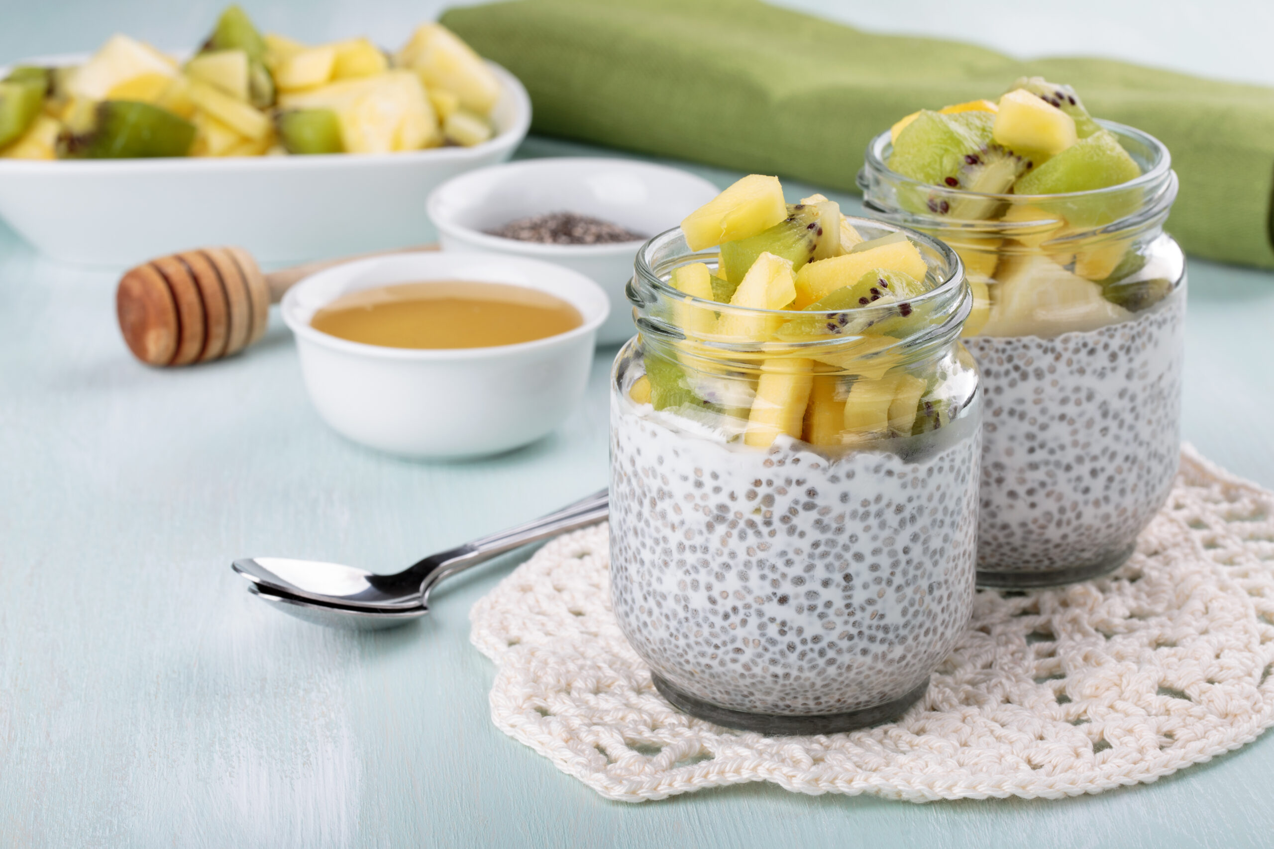 Two glass jars filled with chia pudding, topped with diced kiwi and pineapple pieces. A crochet doily, a spoon, a bowl of fruit, a green cloth, a small bowl of honey, a honey dipper, and a bowl of chia seeds are in the background on a light blue table.