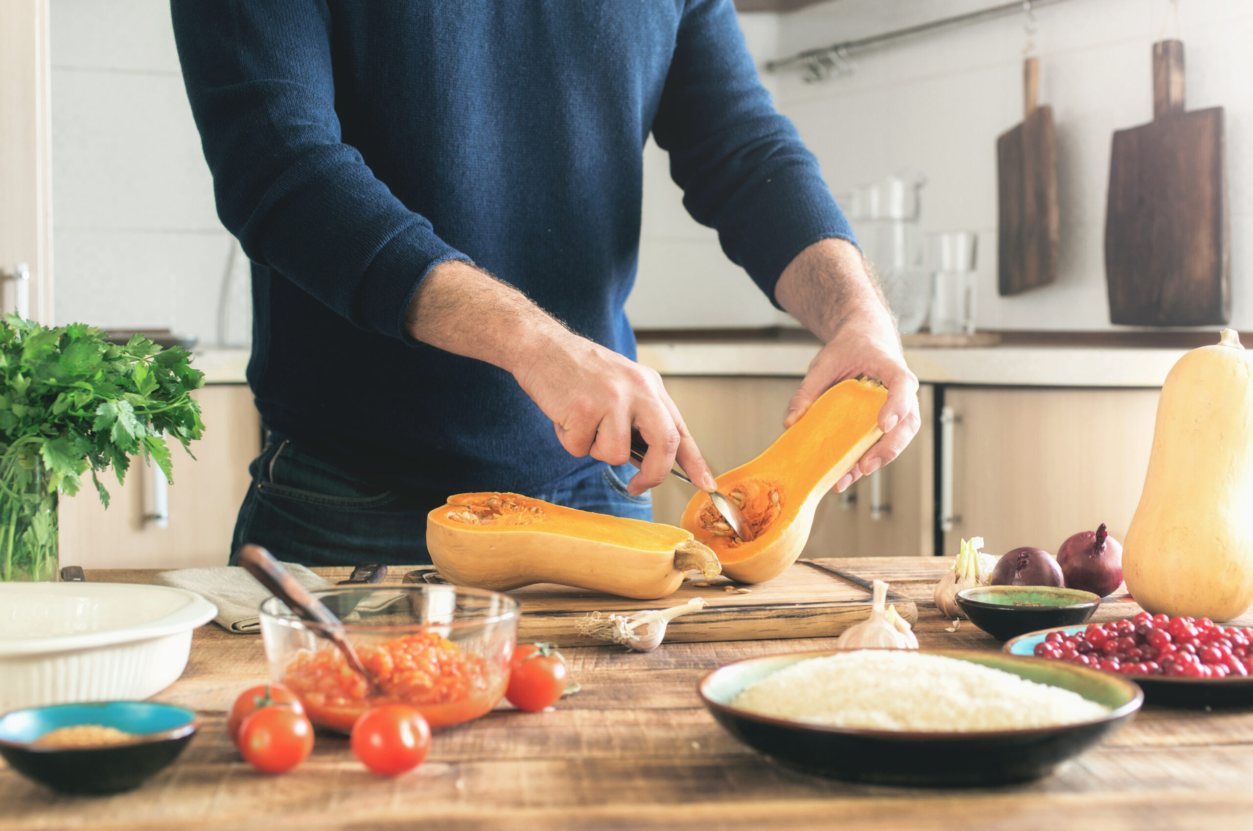 A person in a dark blue sweater is cutting a butternut squash in half on a wooden countertop. The kitchen has a variety of ingredients and utensils, including cherry tomatoes, rice, pomegranate seeds, and fresh herbs.