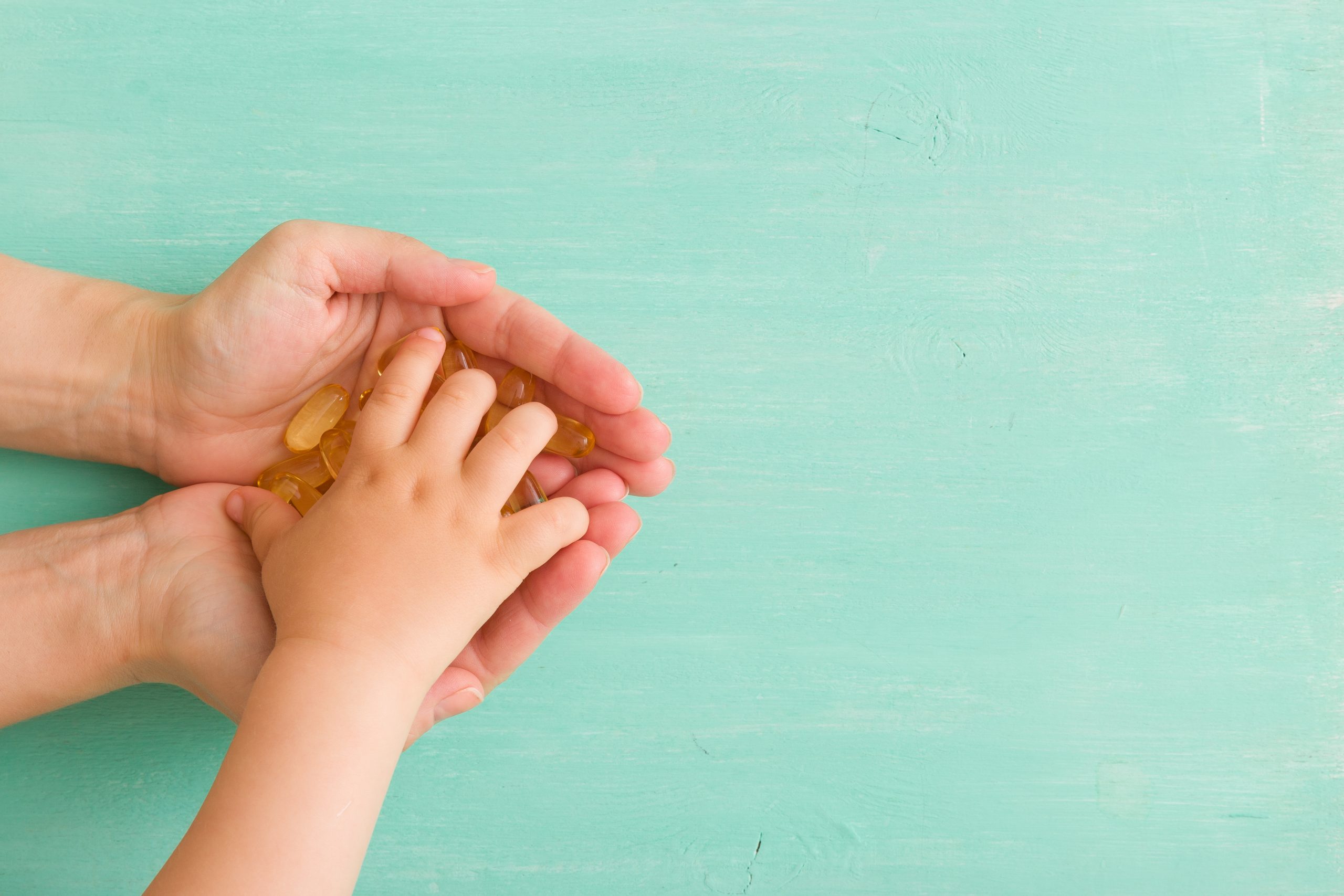 Adult hands holding a child's hand, both grasping a small pile of golden gummy vitamins against a light teal wooden background.