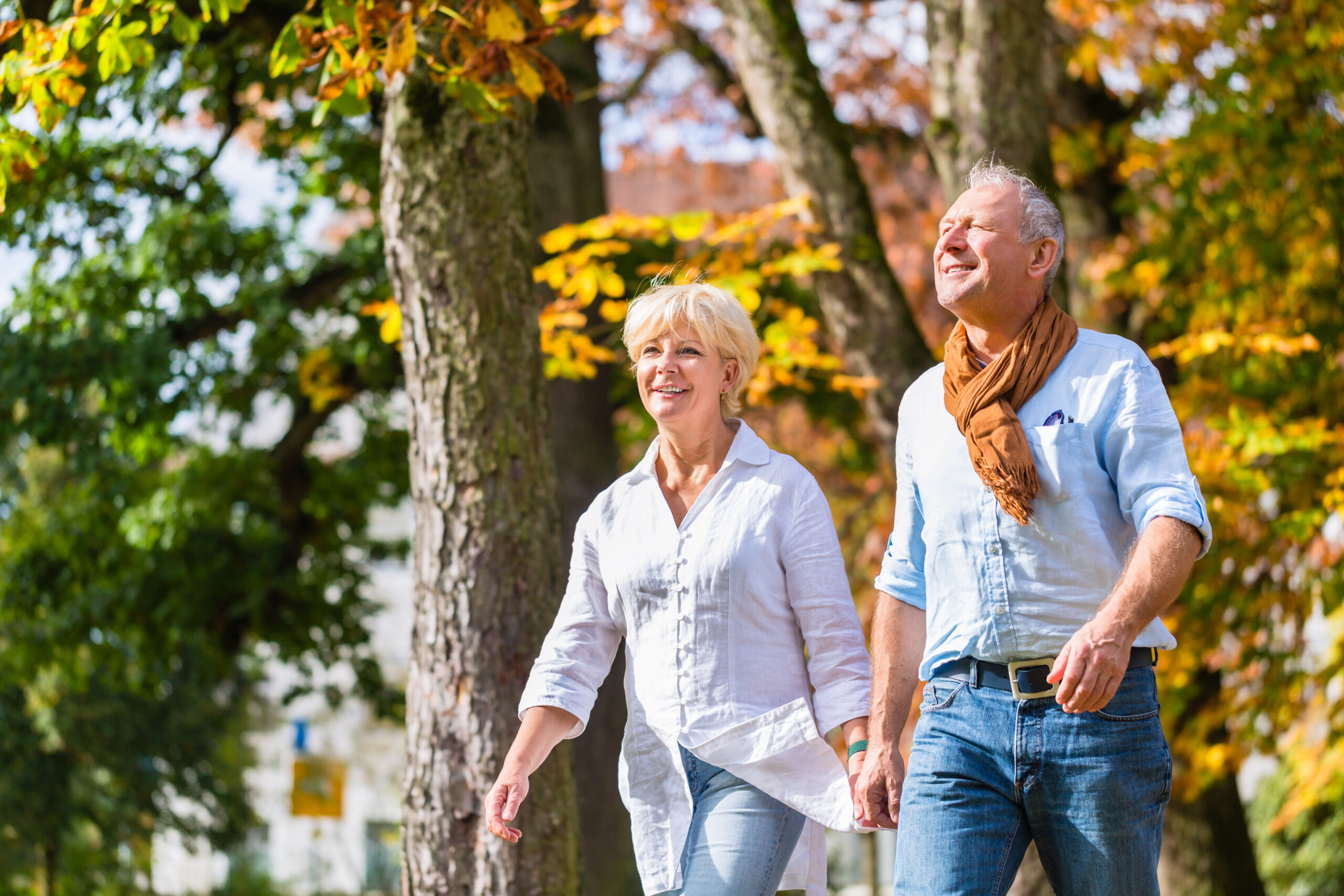 An older couple holding hands and walking in a park on a sunny day. Both are smiling and wearing light-colored shirts, with the man also wearing a scarf. The trees around them have autumn leaves, creating a warm and colorful scene.