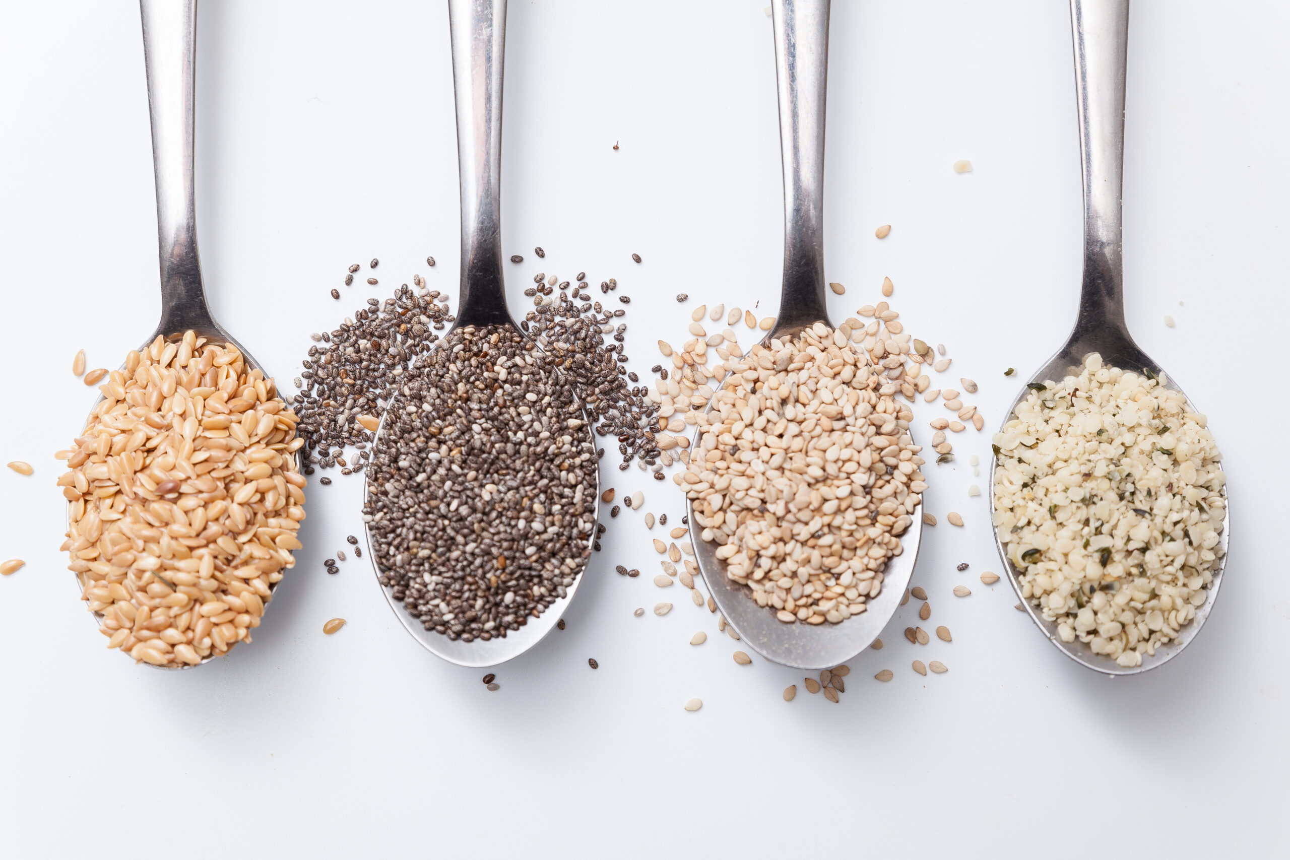 Four metal spoons filled with different types of seeds are lined up against a white background. From left to right: flaxseeds, chia seeds, sesame seeds, and hemp seeds. Some seeds are spilled around the spoons.
