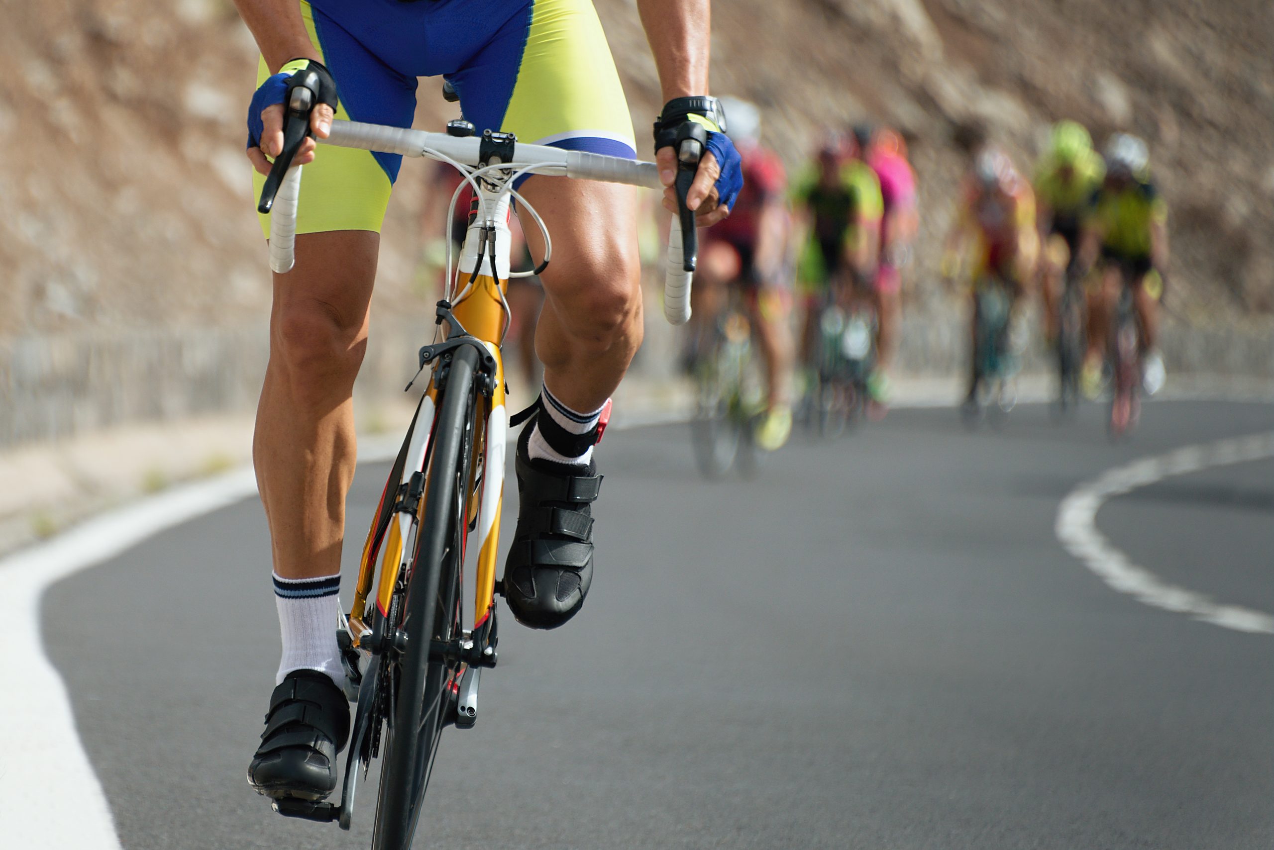 Close-up of a cyclist wearing yellow and blue spandex, black shoes, and gloves, gripping the handlebars while riding on a paved road. A group of cyclists in blurred background also riding on the road, with a rocky, sloped terrain on the left side.