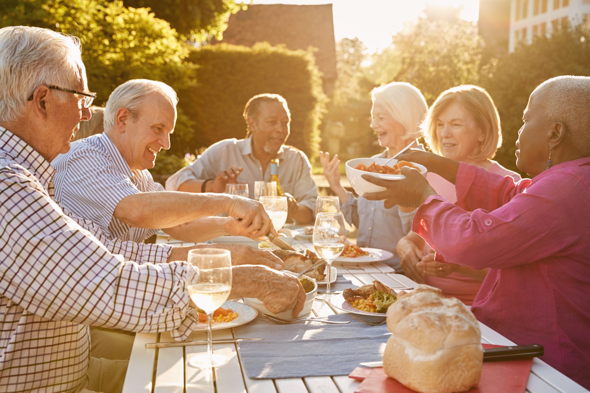 A group of older adults seated around an outdoor table, sharing a meal loaded with foods rich in Omega-3 fatty acids — an undercover love story for the liver. They are smiling and engaging in conversation. The table is set with dishes of food, bread, and glasses of wine under the sunny sky, conveying a warm, social atmosphere.