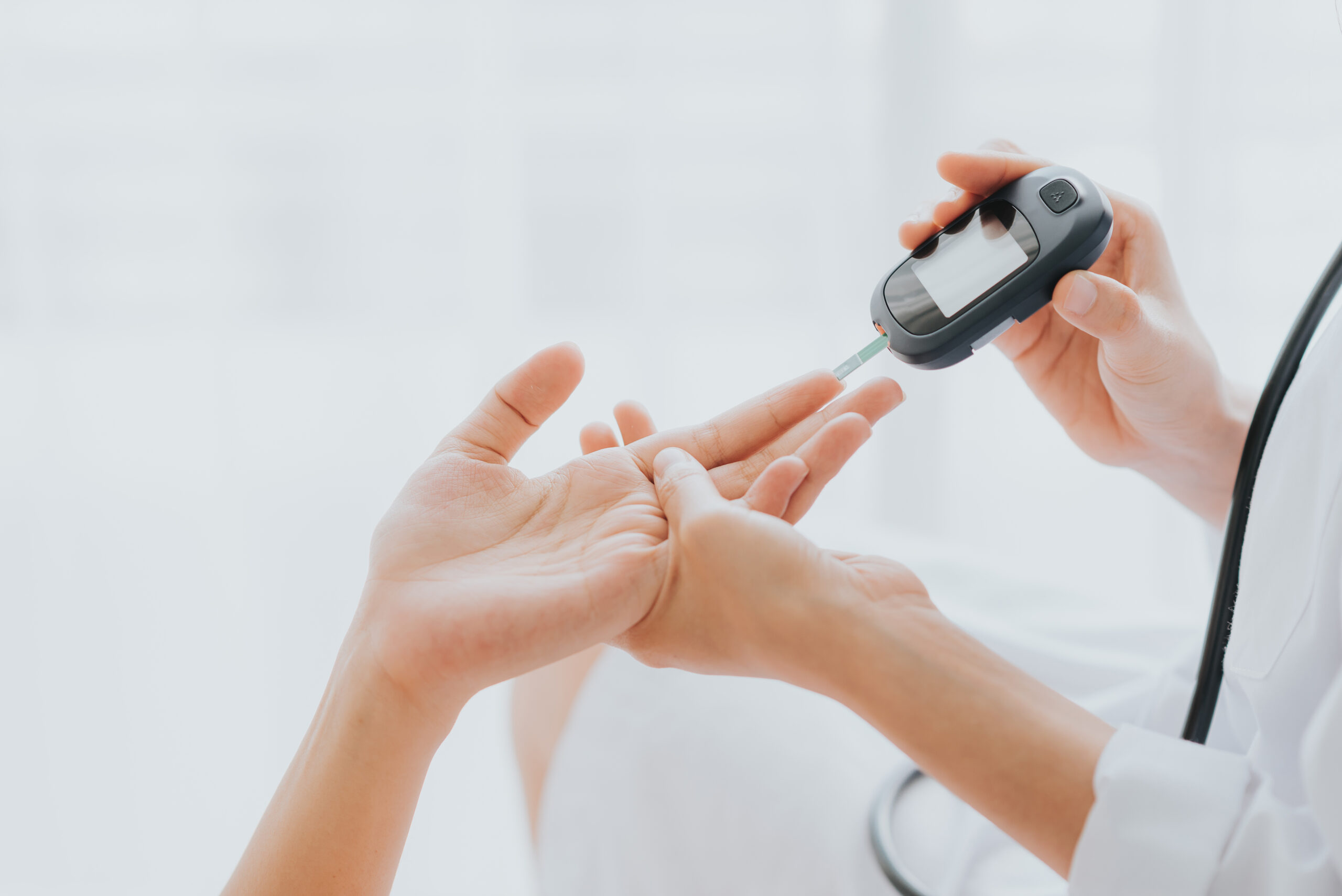 A healthcare professional uses a glucose meter to check blood sugar levels by pricking a patient's finger. The patient’s hand is extended, and the professional holds the meter close to the finger.