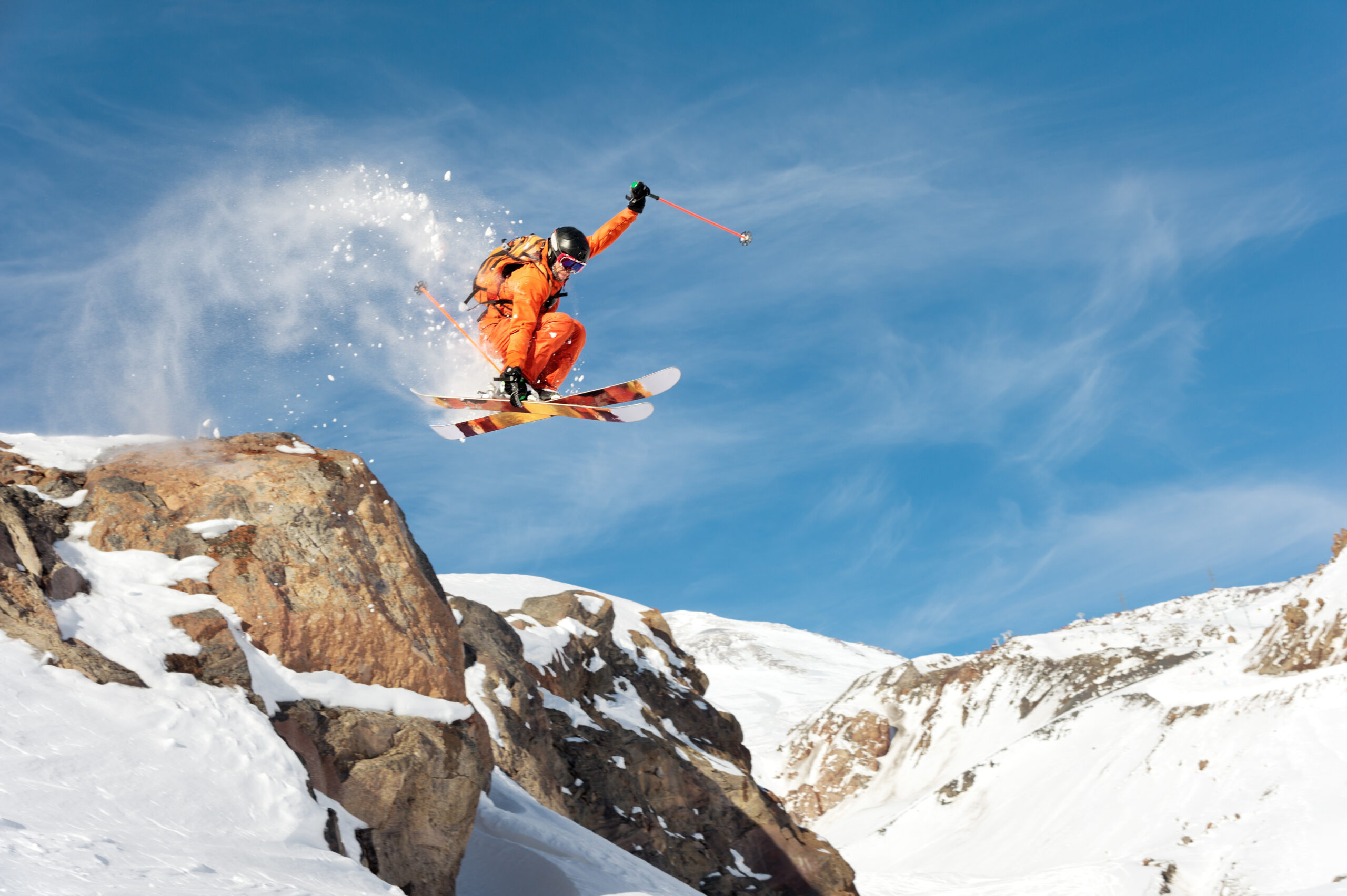 A skier in an orange outfit performs a jump off a rocky ledge on a snowy mountain, with snow spraying around them. The sky is clear and blue, and the mountain landscape is visible in the background.