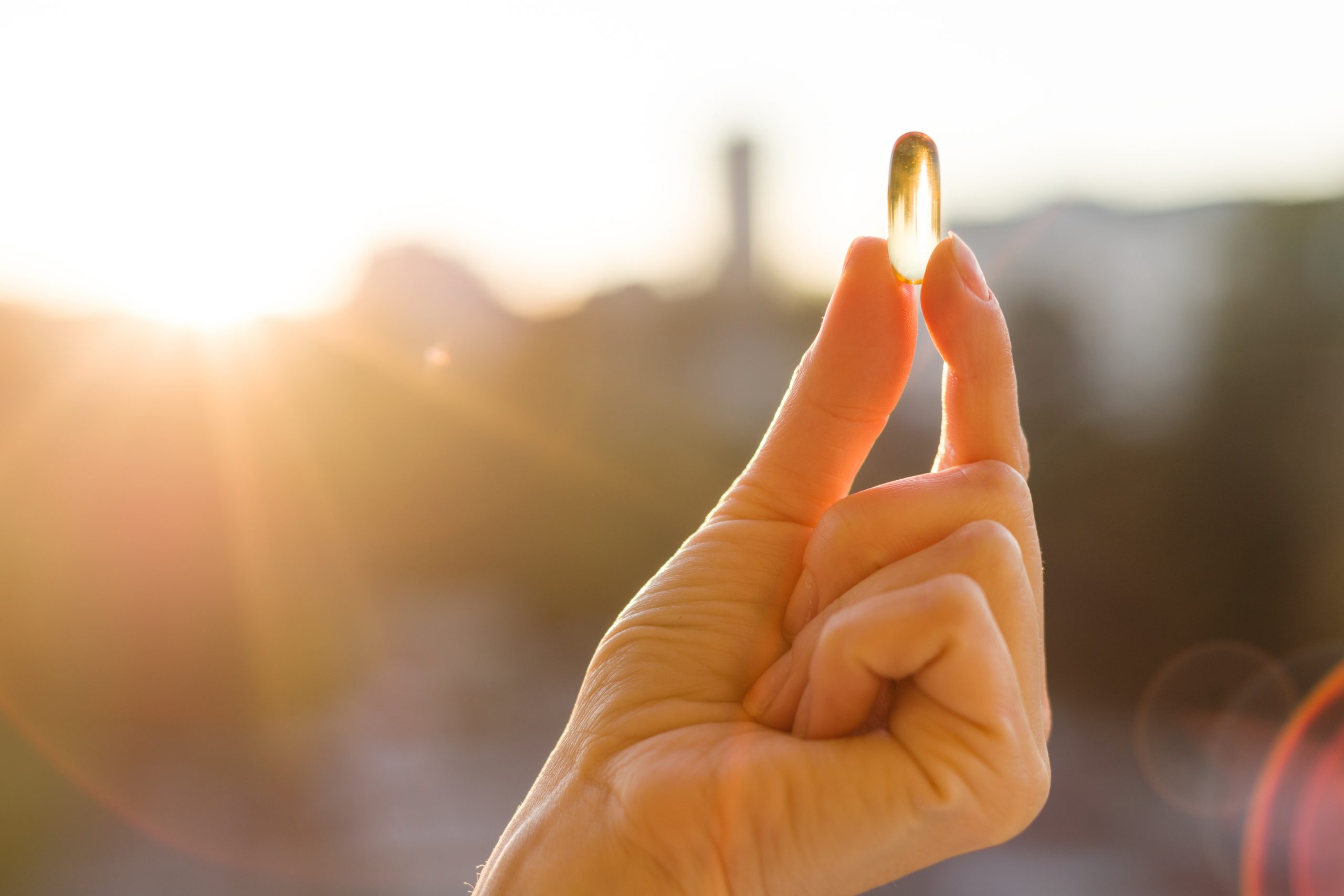 Close-up of a hand holding a transparent yellow capsule between the thumb and index finger, with a blurred background of a bright setting sun and nature.