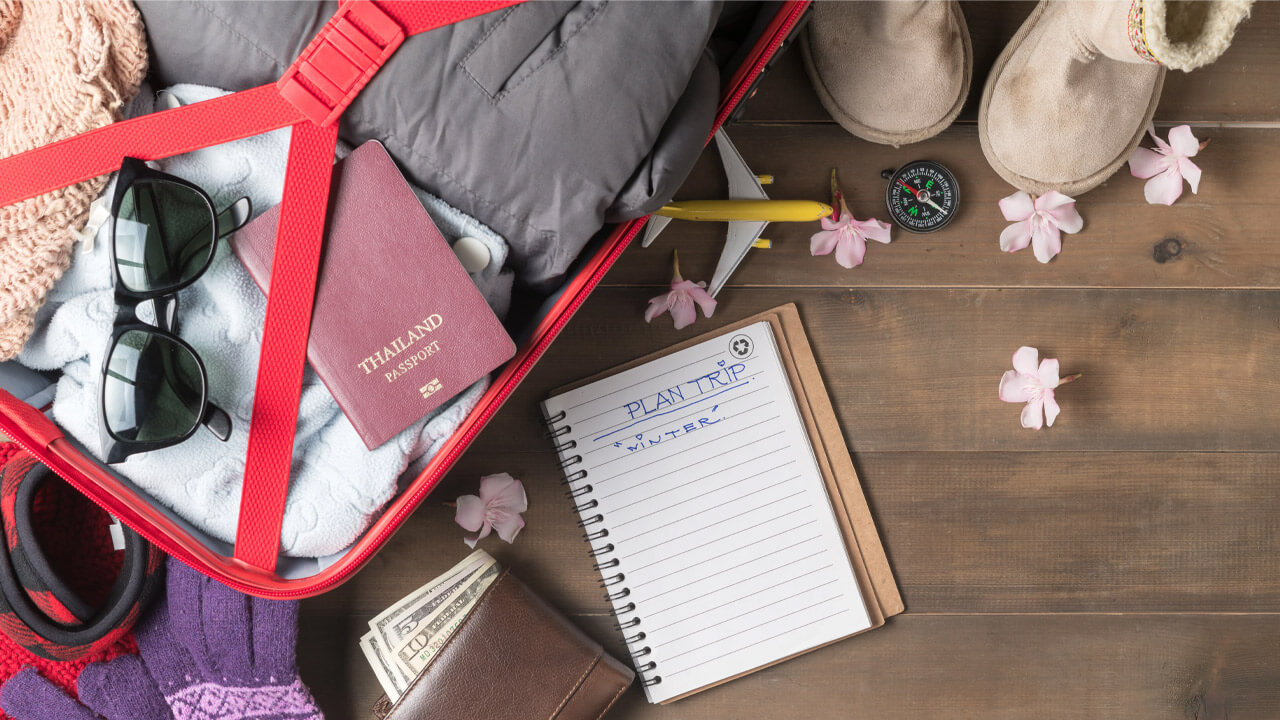 Open red suitcase filled with winter clothes, a passport, sunglasses, and a model airplane lying on the top. Nearby on a wooden floor are boots, a compass, pink flower petals, a wallet with cash, and a notepad with "PLAN TRIP: WINTER" written on it.