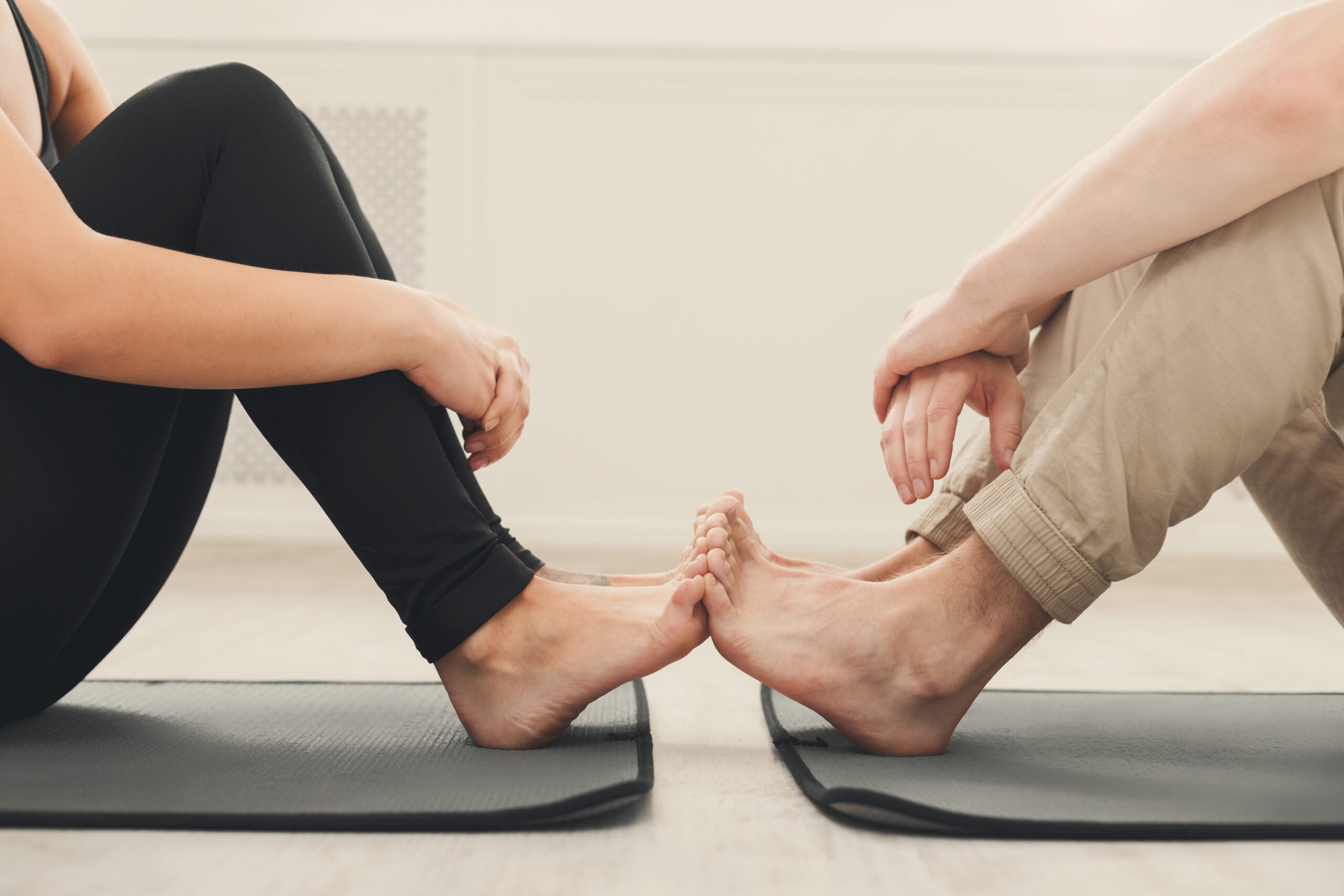 Two individuals sit on yoga mats facing each other, with legs bent and feet touching. One person is wearing black leggings, and the other is wearing beige pants. Their hands are resting on their knees as they engage in a stretching or yoga exercise.