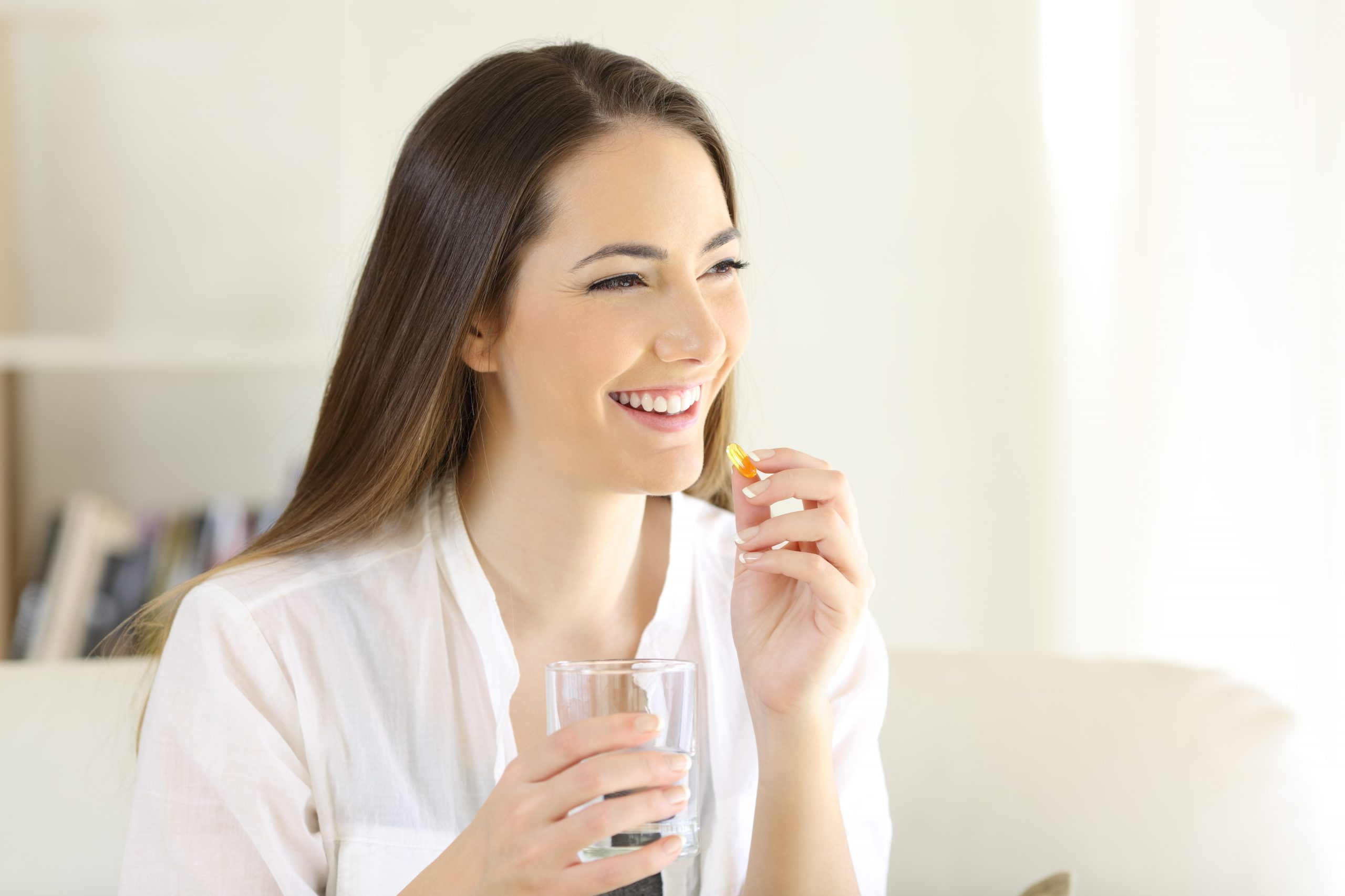 A smiling woman with long brown hair holds a glass of water in one hand and a pill in the other. She is indoors, dressed in a white shirt, and appears to be in a bright, well-lit room.