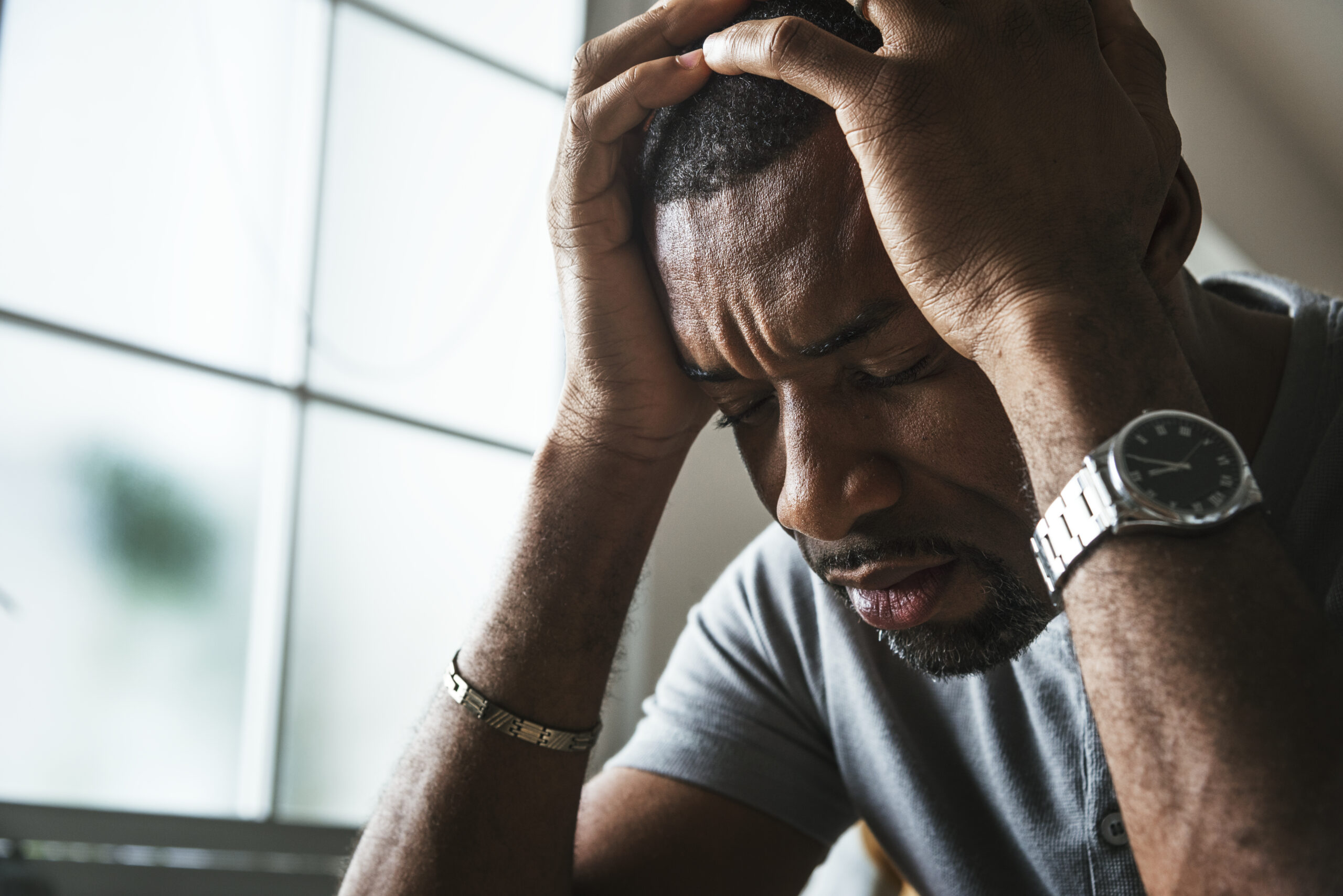 A man sits indoors, leaning his elbows on a surface with his hands clasped over his head. His eyes are closed and his face shows signs of stress or deep thought. He is wearing a short-sleeve shirt, a watch, and a bracelet. A window is visible in the background.