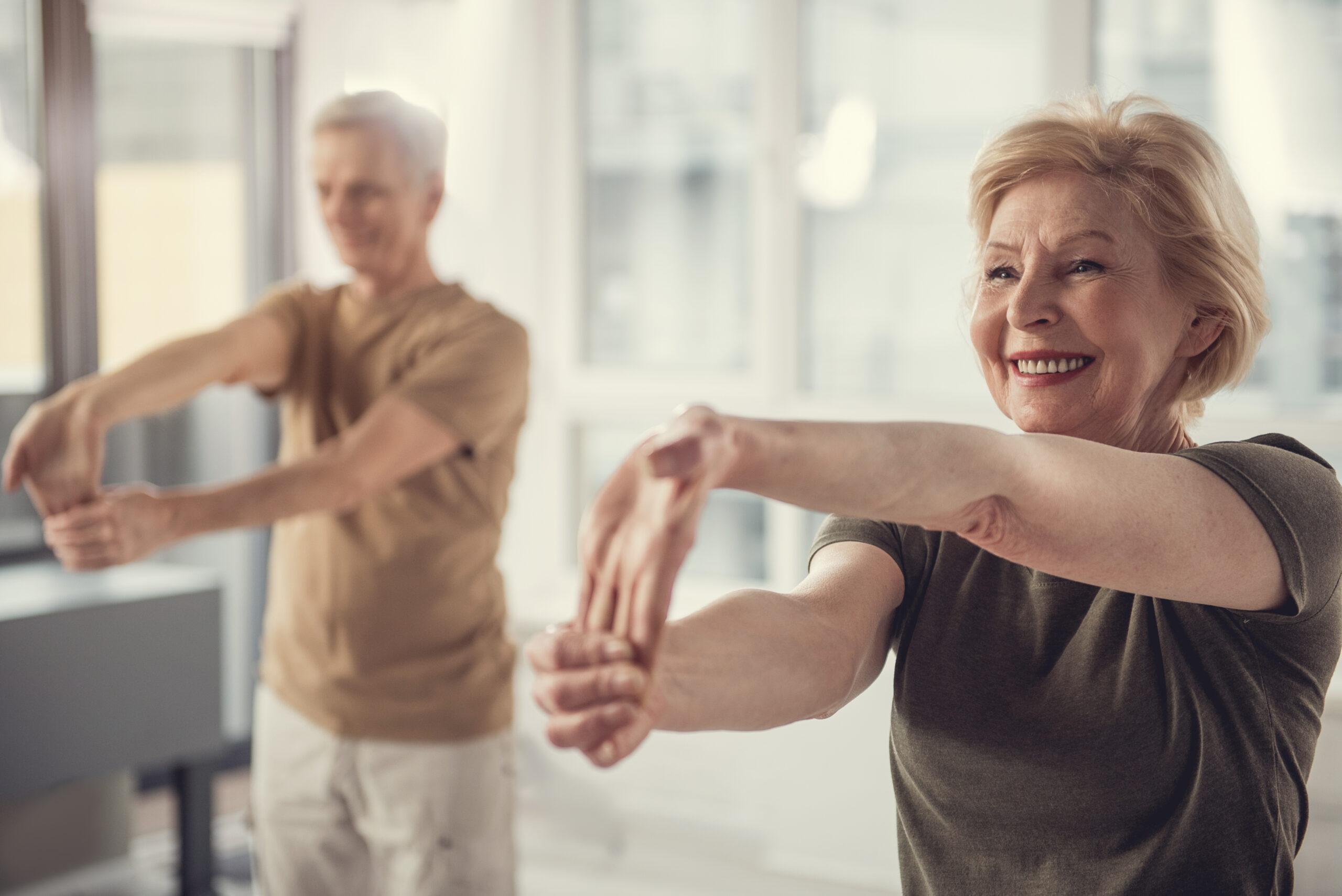Two senior individuals, one man and one woman, are smiling and stretching their arms forward in a bright room. Both appear to be enjoying their exercise routine. The woman is in the foreground, wearing a green shirt, and the man is in the background, wearing a brown shirt.