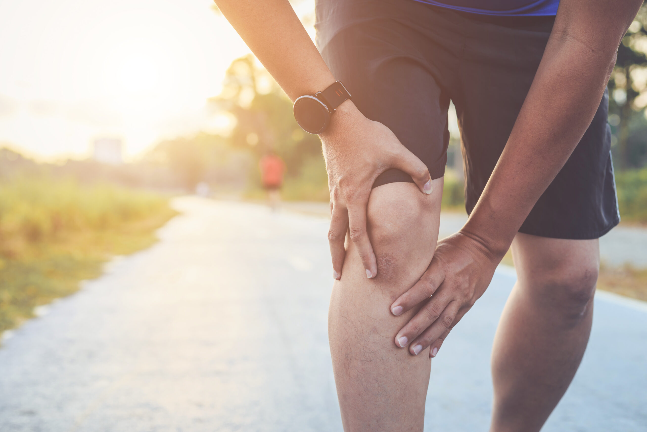 A person wearing a smartwatch is holding their knee in apparent discomfort while standing on an outdoor path. Another person is jogging in the background. The image is taken during daytime with sun flare visible in the top left corner.
