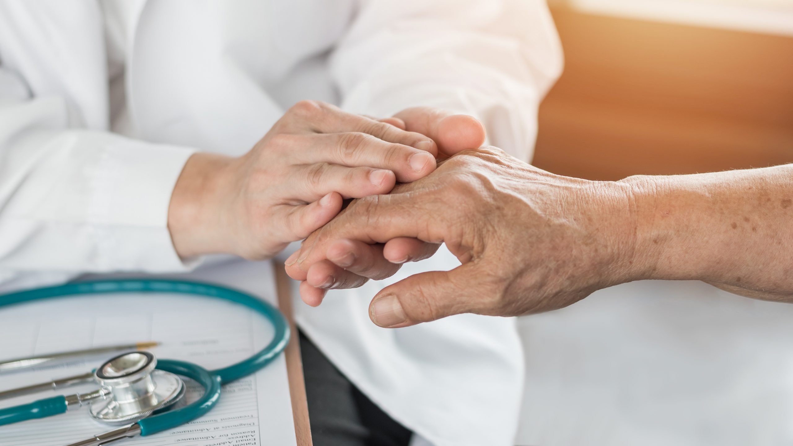 A healthcare professional gently examines an elderly person's hand. The scene includes a stethoscope placed on a clipboard, suggesting a medical setting. The interaction appears compassionate and focused on the patient's well-being.