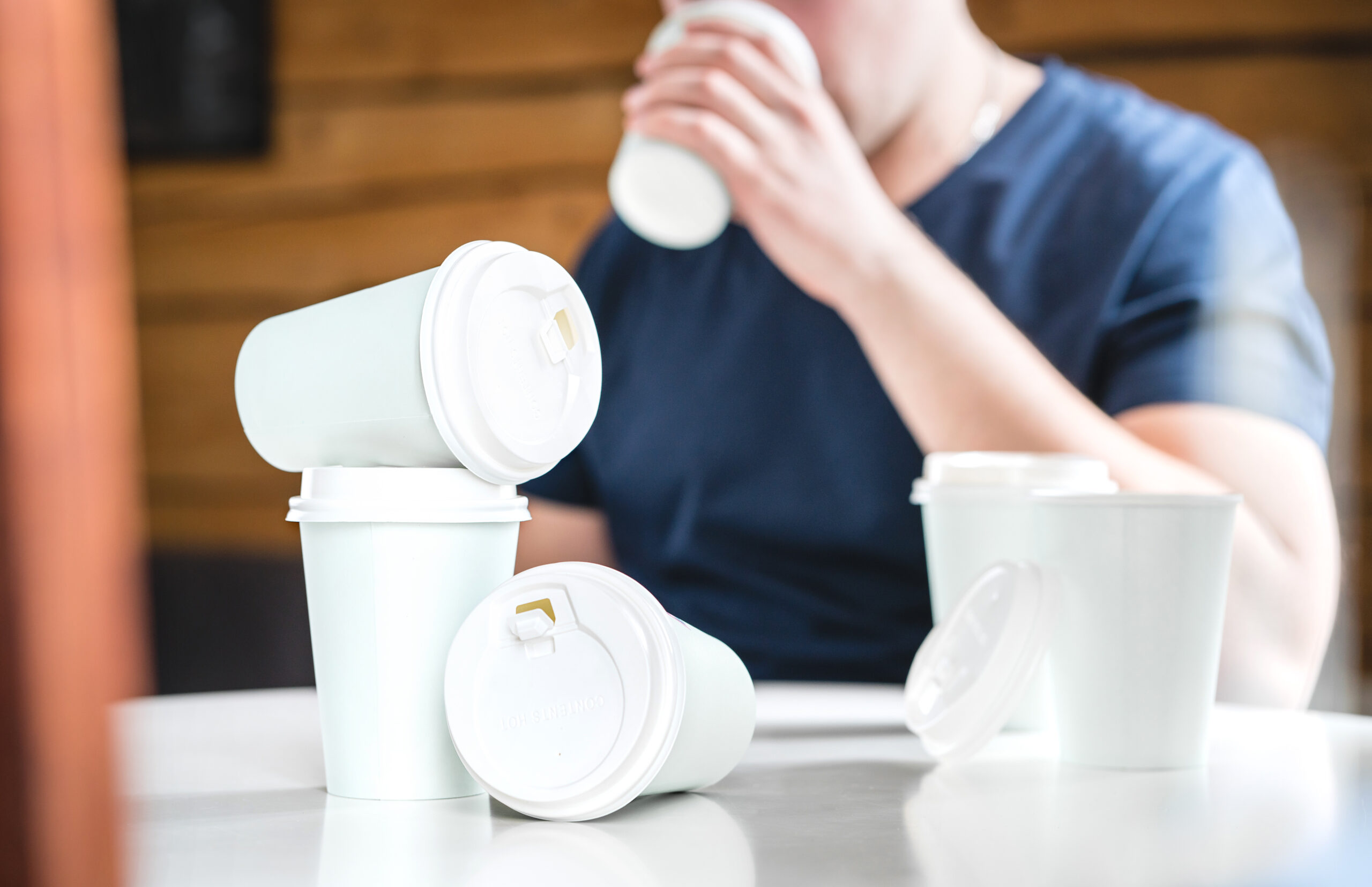 A person in a blue shirt is drinking from a white paper cup with a lid. On the table in front of them are several more white paper cups with lids, two stacked on top of each other and two lying on their sides. The background includes wood paneling.