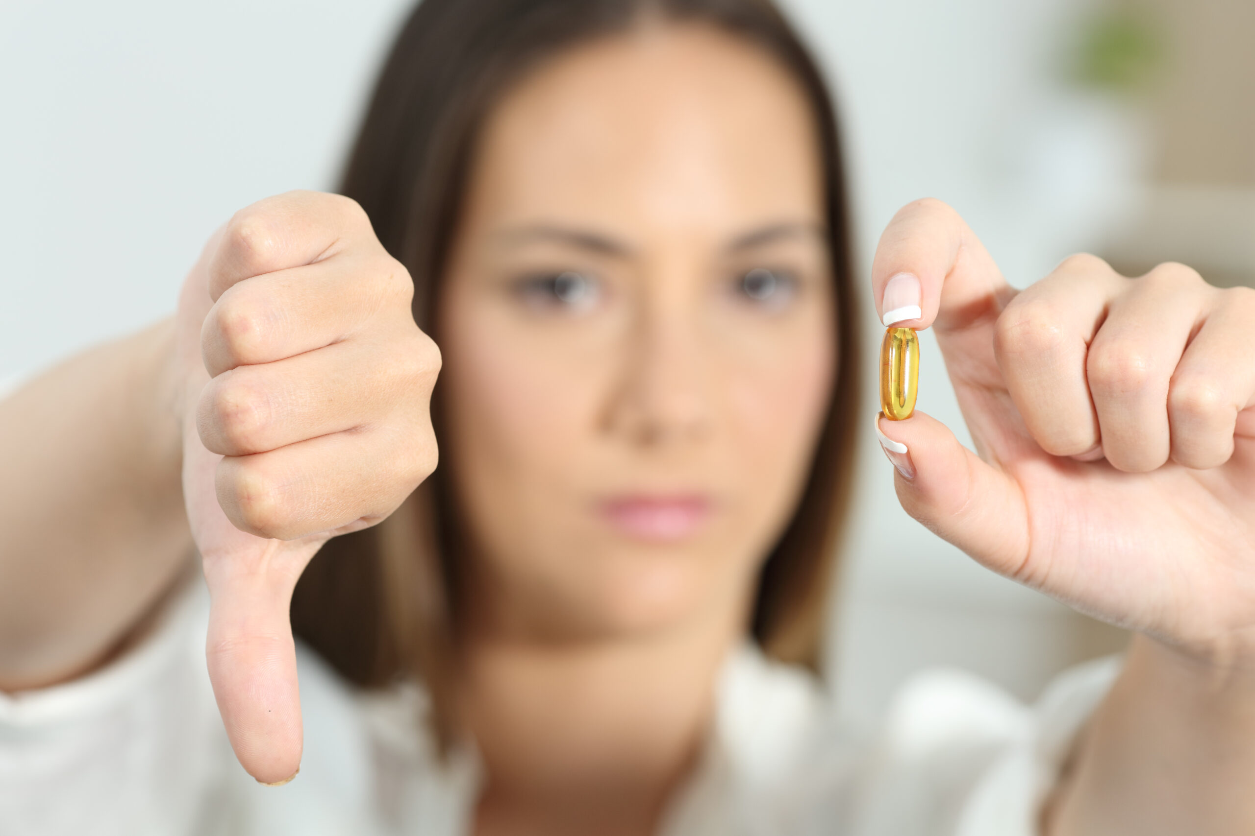 A woman giving a thumbs-down gesture while holding a yellow pill in her other hand. The background is blurred.