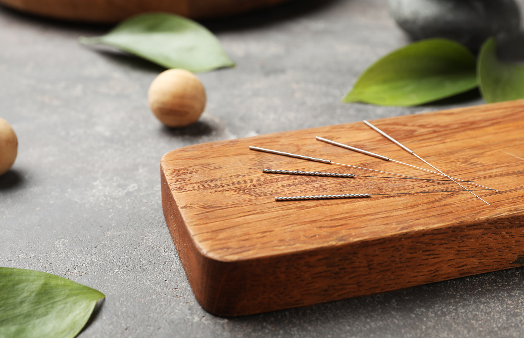 Thin acupuncture needles are neatly arranged on a rectangular wooden board. The board is placed on a gray textured surface, surrounded by green leaves and wooden massage balls, creating a serene and natural spa-like setting.
