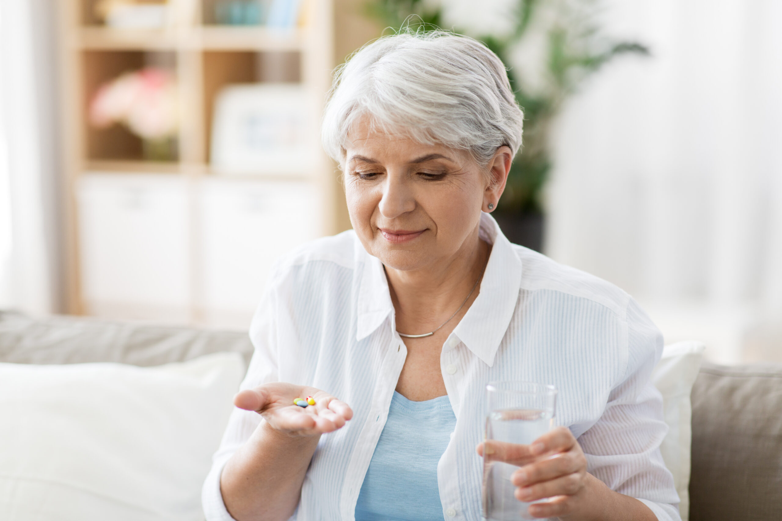 An older woman with short gray hair is sitting on a couch. She is holding a few pills in her left hand and a glass of water in her right hand, preparing to take the medication. There is a bookshelf and some greenery visible in the background.